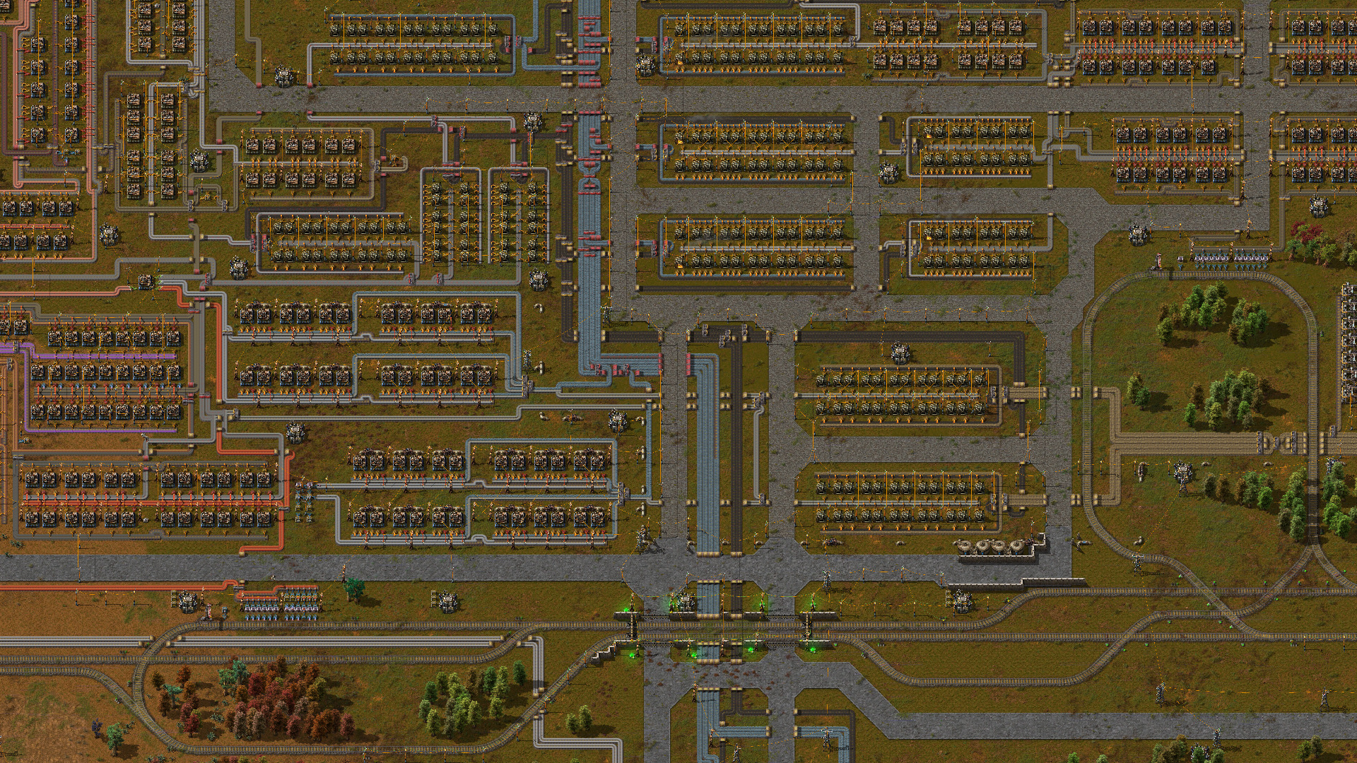 Factorio — A top-down view of an industrial base in Factorio with multiple factory units, interconnected pipelines, rail lines, and surrounding green areas.